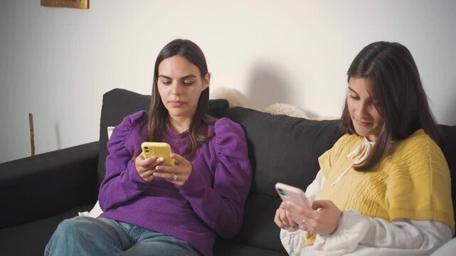Young Latina friends sitting on their sofa distracted and ignoring each other while using their smartphones. Concept of digital distraction, smartphone dependence, social disconnection, lack of commun