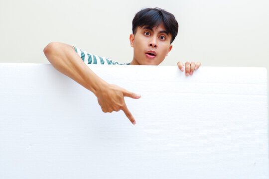 Young Southeast Asian man pointing down at blank white board with funny shocked expression behind wall against plain background ideal for commercial promotion advertising copy space