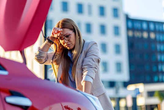 Exhausted business woman with glasses wiping sweat near open engine hood of broken car.