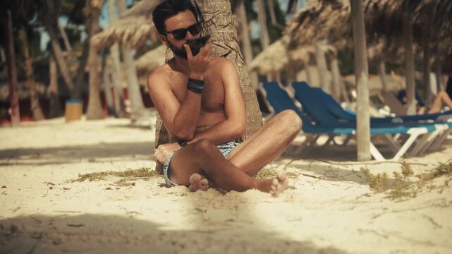 Vacationer Sitting On Sandy Beach Near Palm Tree While Using Smartphone During Summer Holiday