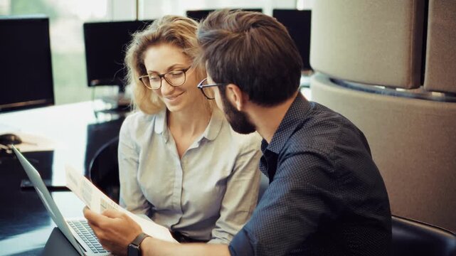 Office Workers Discussing Project Documents While Sitting At Desk With Laptop In Modern Workspace