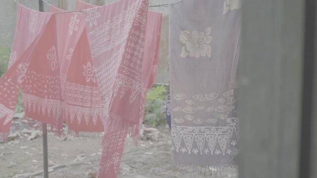 Ungraded log footage of pink and purple batik fabrics with fringed edges hanging on a clothesline and fluttering in strong wind outdoors, traditional Indonesian batik textile