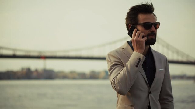 Businessman Talking On Smartphone While Standing Near River With Bridge 