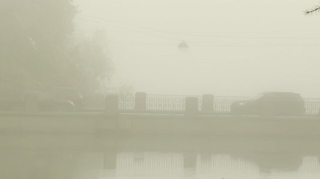 Cars drive silently across a bridge through dense fog over a still canal. The mysterious view creates a quiet mood in Catherine Park, Pushkin, Saint Petersburg, slow motion.