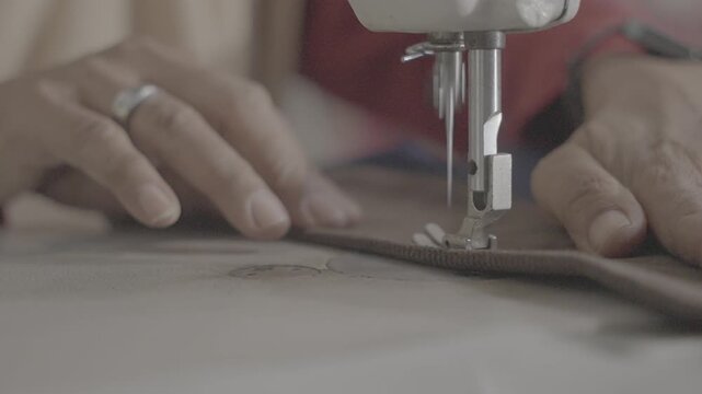 Ungraded log footage of a close up of hands guiding brown fabric under the needle of a sewing machine while stitching, tailoring and garment making craft, flat color profile for color grading