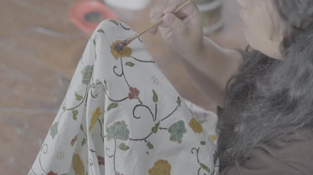 Ungraded log footage of a medium close up of a woman painting colorful floral motifs on white batik fabric with a brush, traditional Indonesian batik coloring