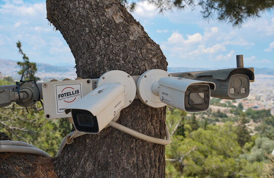Multiple white surveillance cameras mounted on a tree trunk with green landscape and blue sky in Athens, Greece.