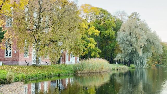A historic red brick pavilion stands beside a serene lake surrounded by autumn trees. The quiet water creates a peaceful and romantic mood. This beautiful scene is at Catherine Park, Pushkin