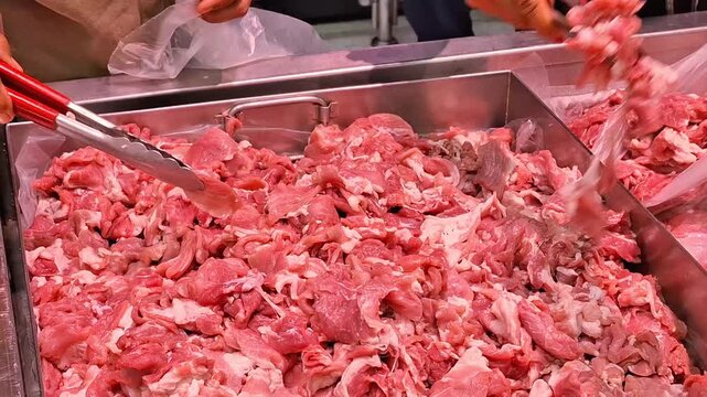 vendor using metal tongs to pick fresh sliced raw pork meat from stainless steel tray at local butcher shop market stall