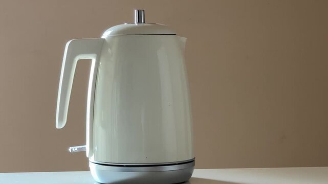 White Modern Electric Kettle Placed on Silver Base by Hand, Switched on, and Heating With Steam Rising, Minimal Indoor Kitchen With Beige Wall, Static Medium Shot at Eye Level, Daylight