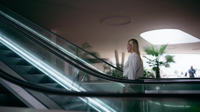 Woman riding modern illuminated escalator upward through contemporary shopping mall, moving from lower level to an upper floor surrounded by neon lights and modern interior architecture