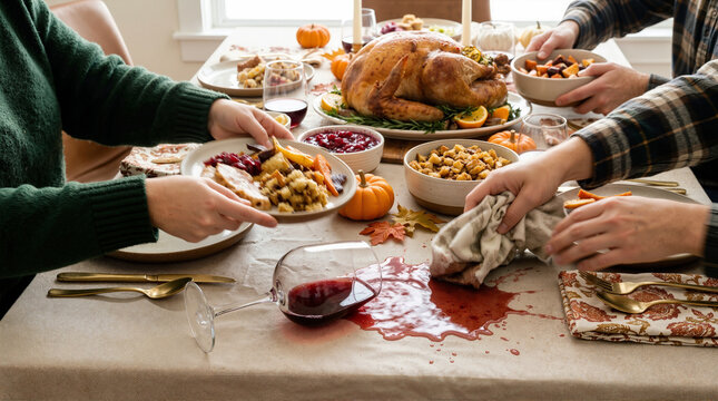 Red wine spilled on table during Thanksgiving dinner. Person wiping accidental spill with napkin while family eats festive meal. Holiday chaos and feast concept