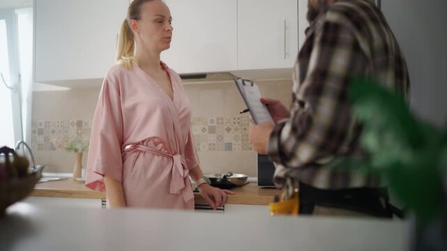 Concerned homeowner in a pink robe discussing kitchen appliance repair options with a professional handyman, who is explaining the job estimate from his clipboard in the home interior