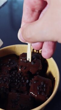 Hand lifts chocolate brownie bite from bowl with wooden pick. Glossy chocolate and small chips make this rich dessert tempting, satisfying sweet craving with indulgent treat, showing piece held on