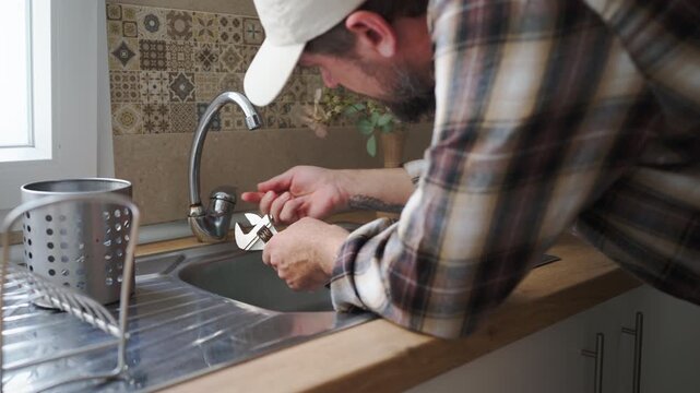 Competent handyman with a beard wearing a plaid shirt and cap, skillfully repairing a leaking faucet in a modern kitchen sink using a specialized plumbing wrench for maintenance