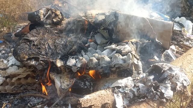 Outdoor burning trash pile with small flames smoke rising, charred debris blackened waste materials smoldering, dry ground sunlight, open fire burning garbage outdoors environmental pollution scene