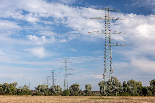 High voltage transmission tower rises at power line construction site with suspended conductors and fencing. Steel lattice structure represents grid expansion and energy engineering under cloudy sky