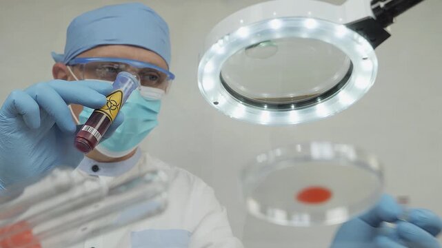 A laboratory technician wearing gloves and safety goggles examines a sample in a vial under a magnifying lamp, focusing on a Petri dish in an indoor lab setting.