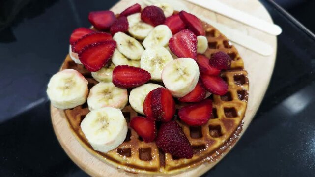 Fruit topped waffle rests on wooden board, offering colorful brunch cafe dish. Sliced strawberries and bananas cover golden grid patterned pastry beside white plate and utensils, ready to serve.