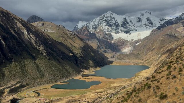 High angle view reveals twin glacial lakes at jahuacocha campsite nestled in deep valley floor beneath massive snowcapped peaks in cordillera huayhuash peruvian andes landscape