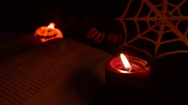 Candle flickers beside open book in dim Halloween setting. Warm light falls across printed pages as pumpkin decor and spiderweb pattern create cozy, spooky reading atmosphere, with flame beside pages.