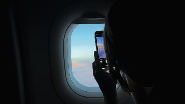 Unrecognizable woman is taking a picture of the sky from an airplane window. The sky is blue and the sun is setting