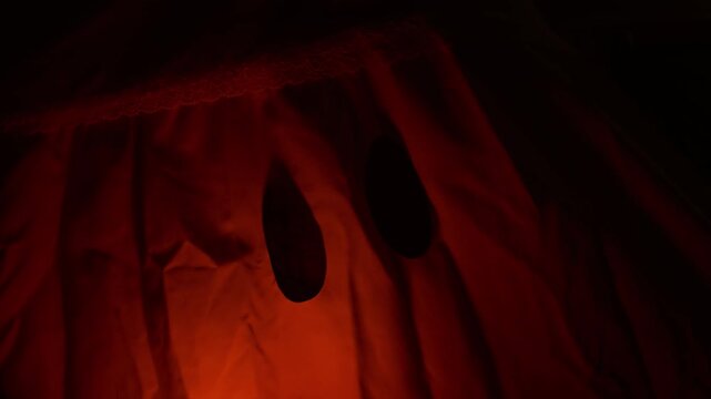 Ghostly figure hangs motionless in red light, wearing pale sheet with two dark eye holes. Shadowy Halloween decoration creates eerie, unsettling mood against nearly black background.