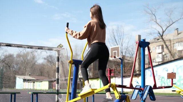 Back view of a young woman doing cardio exercises on an elliptical trainer machine at an outdoor gym, promoting a healthy and active lifestyle on a sunny day in the park