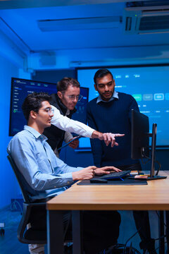 Tech team collaborating on computer software project pointing at screen during discussion in modern control room with blue lighting