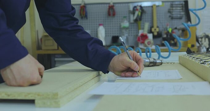 Design and assembly of wooden furniture in a carpentry workshop. A furniture maker in blue workwear measuring a particle board part with a tape measure on a workbench in a manufacturing workshop. 