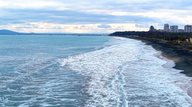 Waves crashing on the shore with city skyline in the background at beachfront. The ocean waves crash onto the sandy shore near the city. People can be seen in the distance.