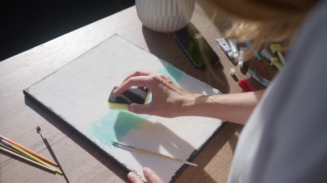 Woman applying light blue paint to a textured canvas with a sponge