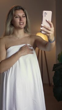 Woman wrapped in a white sheet working as a webcam model during a video call, flirting online and using a smartphone for romantic communication.