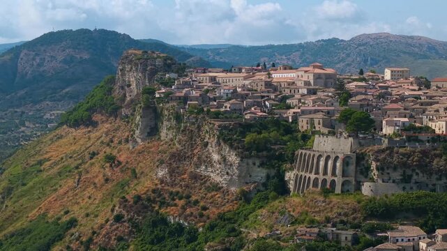 Gerace Calabria Italy 4k drone 70mm view of historic clifftop borgo village. Arched retaining walls on cliff face, telephoto compression with mountain background establishing shot