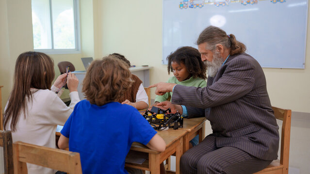 Senior male teacher pointing and guiding diverse multiethnic group of young students assembly robotic kit project in stem class