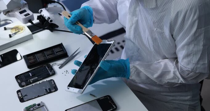Electronics technician in protective clothing cleans a damaged tablet screen with a brush at a repair workstation surrounded by smartphone parts and tools.