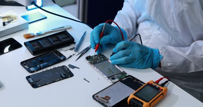 Technician in blue gloves tests an opened smartphone circuit board with probes and a digital multimeter at an electronics repair workbench.