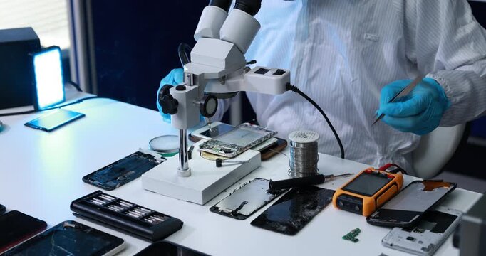 Technician in gloves repairs smartphones and circuit boards under a microscope at an electronics workshop table with tools and spare parts.