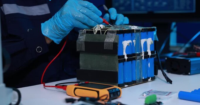 Gloved technician tests a damaged lithium battery pack with probes and a digital meter on a workshop table.