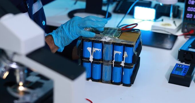 Technician wearing blue gloves examines a damaged lithium battery pack with exposed cells and wires in an electronics repair lab.