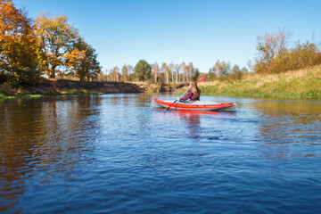 A person paddling a red inflatable kayak on a wide river along an autumn forest shore © WoodHunt