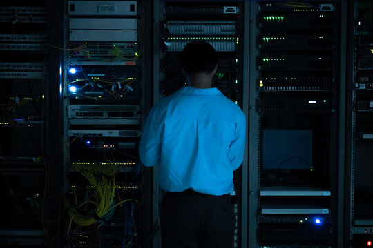 Focused Asian male IT network administrator uses a laptop to monitor server performance inside a dark data center room managing complex corporate cybersecurity operations and infrastructure.