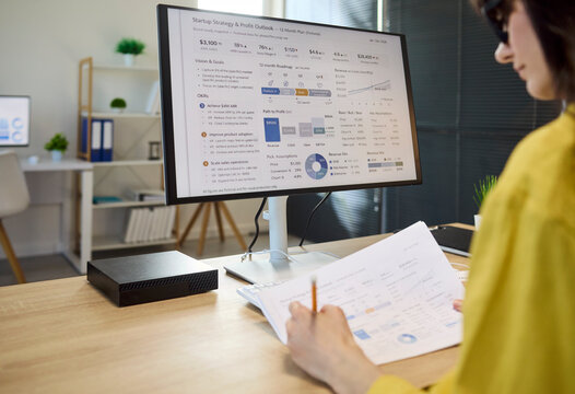 Woman accountant performs finance analysis on computer. She reviews charts on screen and paperwork, turning data into reports for planning forecasts. Modern finance analytics concept in office.