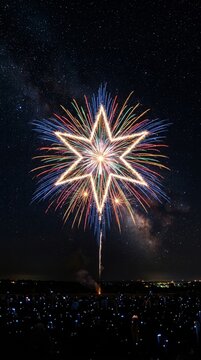 Star shaped firework exploding in the night sky over a silhouette of a crowd holding glowing lights in an outdoor field