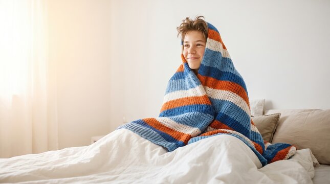 Young boy sits comfortably in bed while wrapped in a soft colorful striped blanket to stay cozy during a bright morning at home and smiles while looking away from the camera