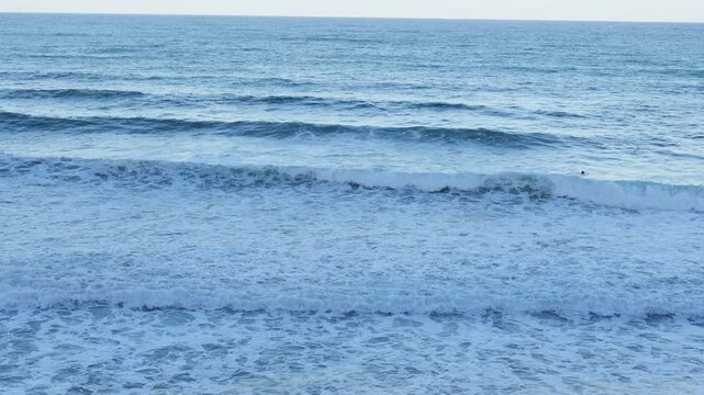 High angle background perspective of wide blue sea surface with gentle rolling white foam waves and water ripples on sunny summer day along coastal shore