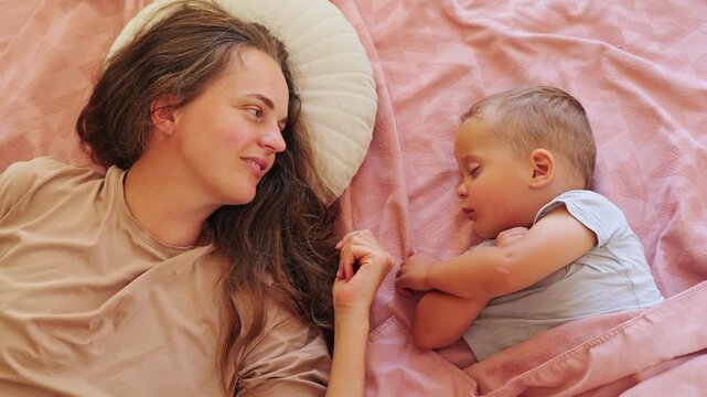 Loving mother lying next to her sleeping toddler boy in cozy bed looking at him with smile during quiet daytime nap family love concept