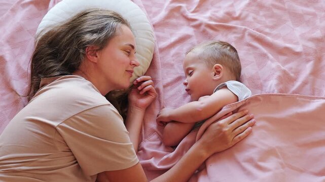 Caring young mother covering sleeping toddler boy with soft pink blanket lying together in cozy bed at home during daytime nap