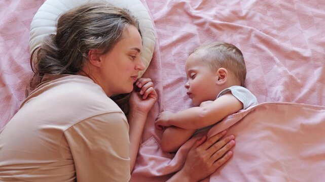 Loving mother watching her little toddler son sleeping peacefully under pink blanket in cozy bedroom during quiet afternoon nap time
