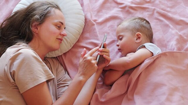 Smiling young mother lying in bed taking picture or video using smartphone while her little toddler son sleeps peacefully beside her